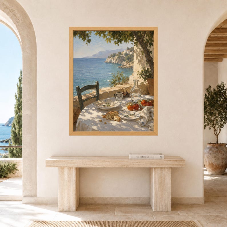 May include: A framed wall art print depicting a coastal scene with a round table set for a meal on a stone terrace. The table features a white cloth, plates with bread, red tomatoes, a glass carafe, and a dark wooden chair under tree shade.