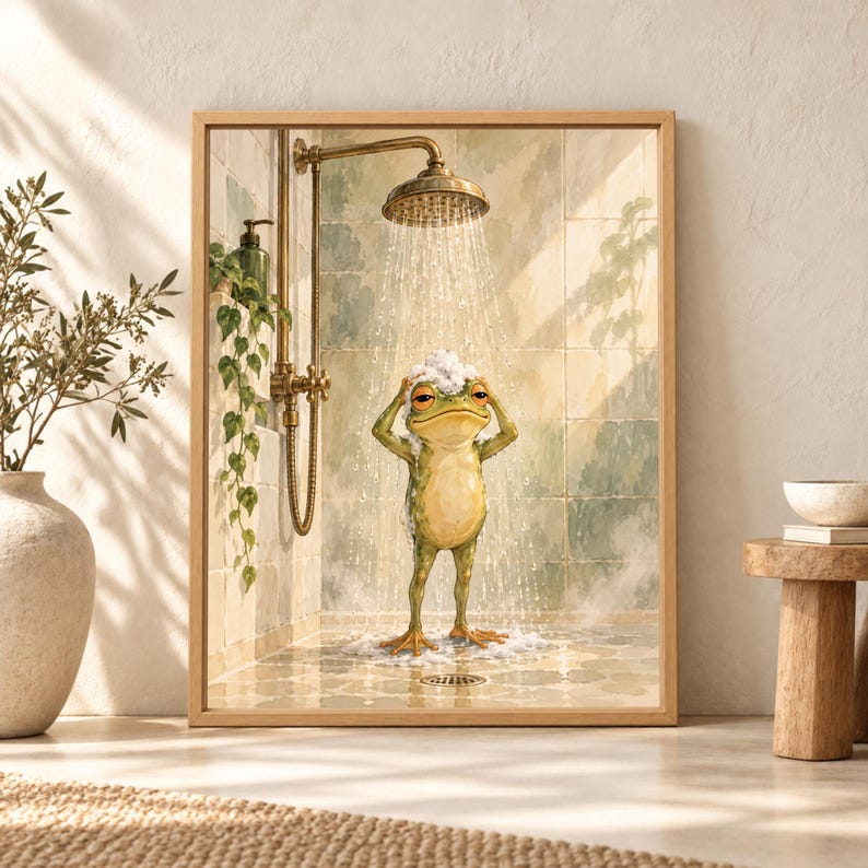 Framed funny frog bathroom wall art showing a green frog washing its head beneath a brass shower with soap bubbles, falling water and soft sage green tiled walls.