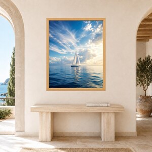Pode incluir: Uma impress&atilde;o de arte de parede retangular apresentando um veleiro branco em um oceano calmo e azul profundo sob um c&eacute;u brilhante com nuvens finas e luz solar dourada. A obra est&aacute; em uma moldura de madeira clara em uma parede neutra.