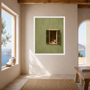 May include: A framed wall art print featuring a sage green wall with an open window. The window has dark green shutters and shows a person sitting on the ledge reading a book. The artwork is displayed in a white frame on a neutral wall.