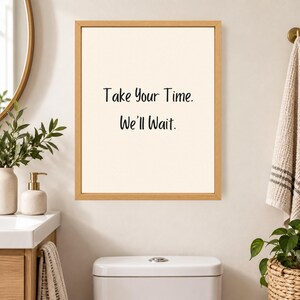May include: A rectangular wall art piece in a light wood frame featuring a cream-colored background with the black text Take Your Time. We'll Wait. centered in the middle. The print is displayed on a wall above a white toilet in a bathroom setting.