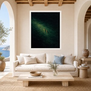 May include: A framed wall art print featuring an aerial view of dark, deep green ocean water with two small figures swimming in a patch of sunlight. The artwork is displayed in a white frame hanging on a beige wall above a cream-colored sofa.