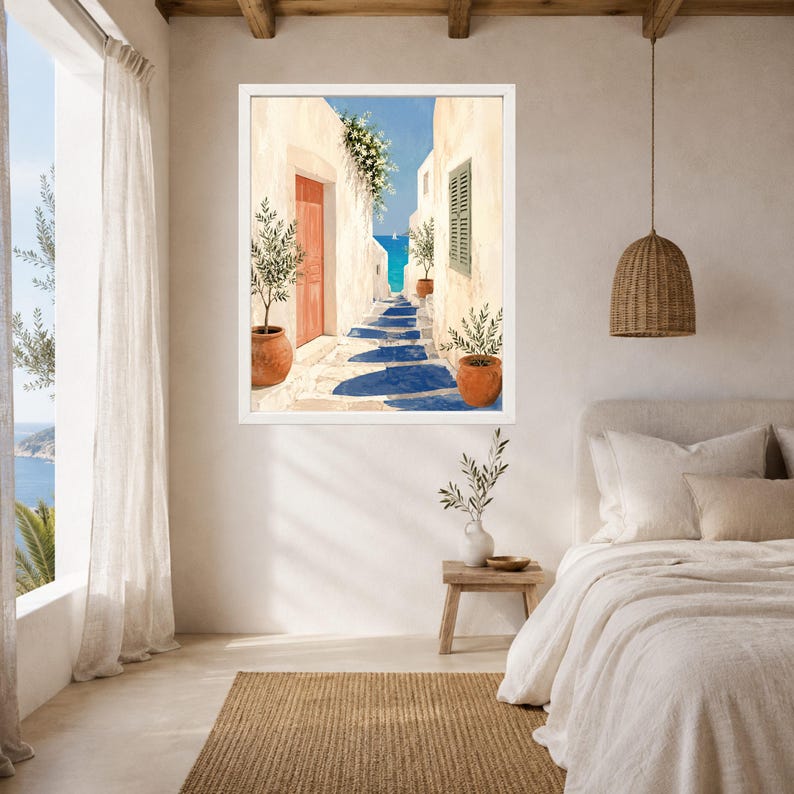 May include: A framed wall art print depicting a Mediterranean coastal alleyway with white stucco buildings, terracotta potted olive trees, a bright orange door, and a view of the blue sea under a clear sky, displayed in a bedroom interior setting.