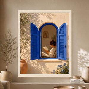 Framed Mediterranean reading wall art showing a woman reading inside an arched window with vivid blue shutters, warm cream plaster walls, olive branches and soft afternoon sunlight.