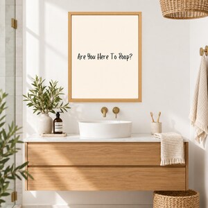 May include: A rectangular wall-mounted sign with a light beige background and a thin wooden frame. The text "Are You Here To Poop?" is printed in black, sans-serif lettering in the center of the sign, which is displayed above a bathroom sink.