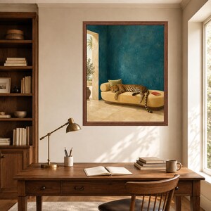 May include: A framed wall art print featuring a leopard resting on a yellow chaise lounge in a room with textured teal walls and a stone floor. The artwork is displayed on a wall above a wooden desk with a lamp, books, and a chair in the foreground.
