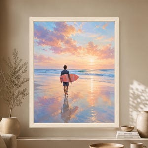 Framed coastal wall art showing a surfer carrying a pink surfboard toward ocean waves at sunset, with peach and lavender clouds reflected across the wet beach.