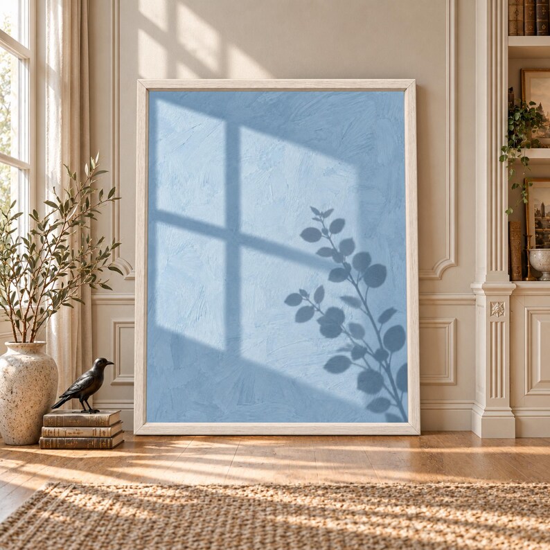 Framed dusty blue botanical wall art showing a soft leafy branch silhouette and large window shadow across a textured pale blue wall in natural sunlight.