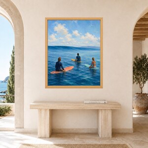 May include: A framed wall art print depicting three individuals sitting on surfboards in the ocean, facing a large wave under a blue sky with white clouds. The surfers are positioned on orange, yellow, and light blue boards in deep blue water.