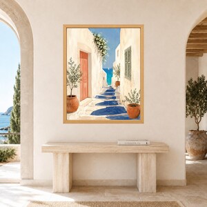 May include: A framed wall art print depicting a Mediterranean coastal alleyway with white stone walls, a bright orange door, green potted olive trees, and a view of the blue sea in the distance. The scene features deep blue shadows on the ground.