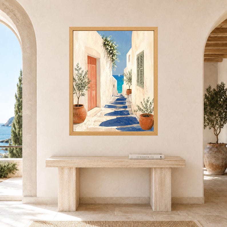 May include: A framed wall art print depicting a Mediterranean coastal alleyway with white stone walls, a bright orange door, green potted olive trees, and a view of the blue sea in the distance. The scene features deep blue shadows on the ground.