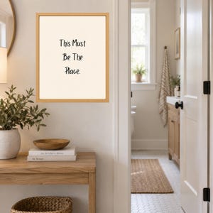 May include: A rectangular wall art piece in a light wood frame featuring a cream-colored background with the black text "This Must Be The Place." The artwork is displayed on a wall above a wooden console table with books and a bowl.