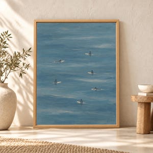 Framed minimal surfer wall art showing six tiny surfers sitting and paddling on cream surfboards across a wide expanse of calm muted blue ocean water.