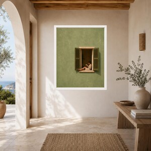 May include: A framed wall art print featuring a sage green wall with an open window. The window has dark green shutters and shows a person sitting on the ledge reading a book. The artwork is displayed in a white frame on a neutral wall.
