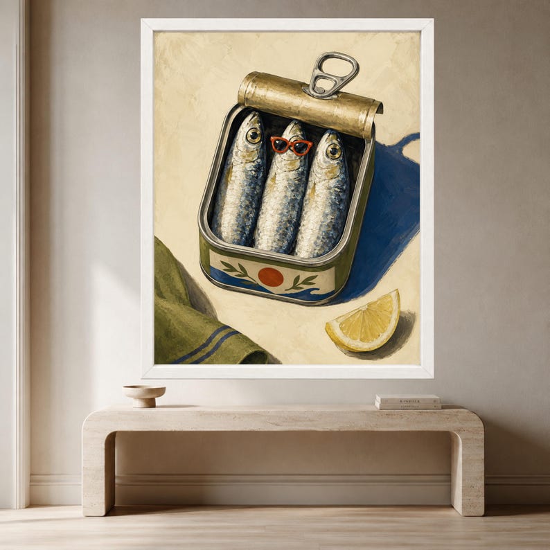 May include: A framed wall art print depicting an open metal tin of three sardines. The middle sardine wears small red-rimmed sunglasses. The tin features a blue and orange graphic design. A slice of yellow lemon sits beside the tin on a light surface.