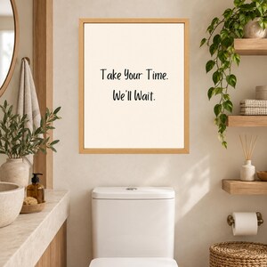 May include: A rectangular wall art piece in a light wood frame featuring a cream-colored background with the black text "Take Your Time. We'll Wait." centered on the print. The artwork is displayed on a wall above a white toilet in a bathroom.