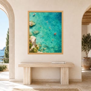 Puede incluir: Una impresi&oacute;n de arte mural rectangular en un marco de madera clara que muestra una vista a&eacute;rea de agua de mar turquesa con acantilados rocosos, un peque&ntilde;o bote blanco y varias personas nadando, incluida una con un flotador rojo.
