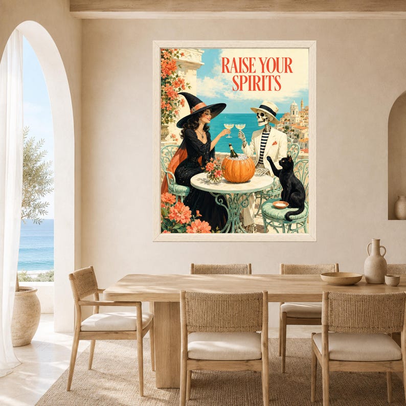 May include: A framed wall art print titled RAISE YOUR SPIRITS featuring a witch in a black dress and a skeleton in a white suit sitting at a table with a pumpkin, wine glasses, and a black cat against a coastal Mediterranean background.