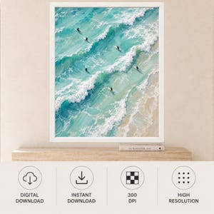 May include: A framed wall art print featuring an aerial view of turquoise ocean waves crashing onto a sandy beach. Several surfers on surfboards are positioned in the water, riding the waves. The painting style uses visible brushstrokes.