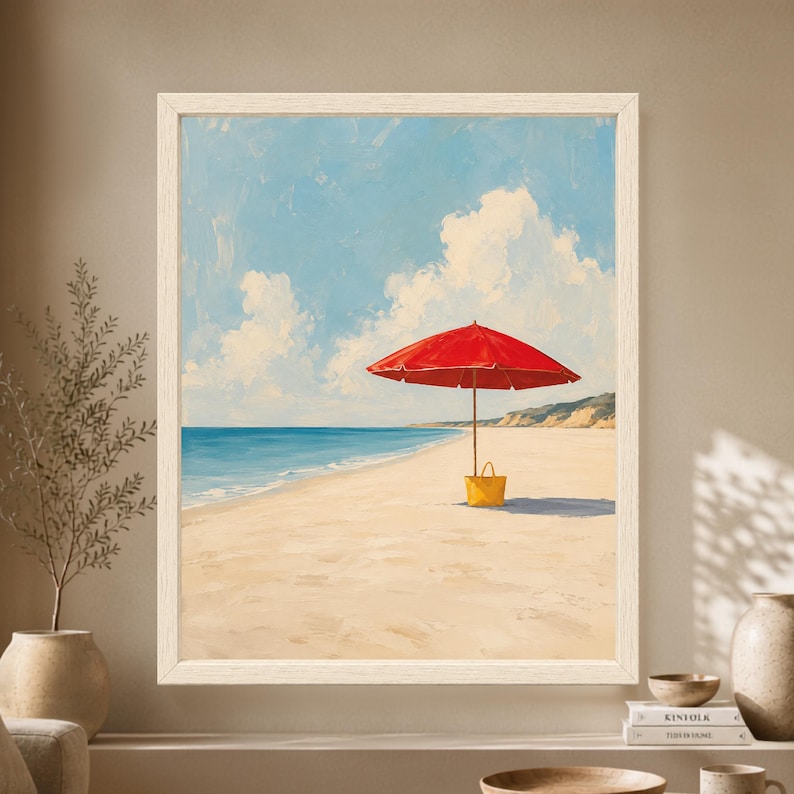 Framed coastal beach wall art showing a single bright red umbrella and yellow beach bag on pale sand beside a calm blue ocean beneath large white summer clouds.