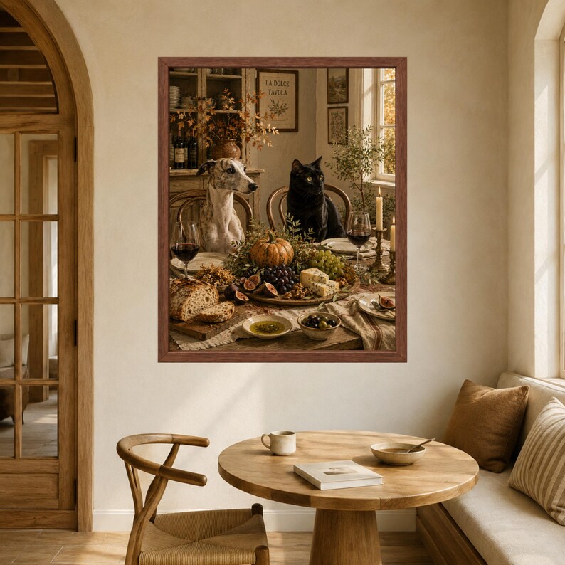 Op de afbeelding: Een ingelijste kunstdruk met een grijs-witte whippet en een zwarte kat aan een eettafel gedekt met een pompoen, druiven, vijgen, kaas, brood, olijven en wijnglazen. De sc&egrave;ne speelt zich af in een rustiek interieur van een eetkamer.