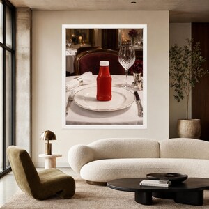 May include: A bright red plastic squeeze bottle of ketchup sits upright in the center of a white ceramic dinner plate. The plate is placed on a white tablecloth, accompanied by a silver fork, a silver knife, and a clear crystal wine glass.