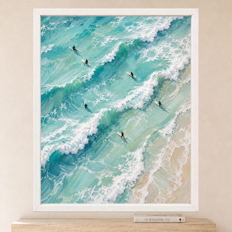 May include: An oil painting in a white frame depicting an aerial view of turquoise ocean waves crashing onto a sandy beach. Several surfers on surfboards are positioned in the water, navigating the rolling white-capped waves and clear blue sea.