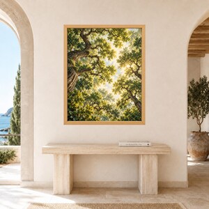 May include: A rectangular wall art print in a light wood frame featuring a low-angle view of a dense forest canopy. The image displays textured tree trunks and lush green leaves with sunlight filtering through the branches toward a bright sky.