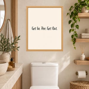 May include: A rectangular wall art print in a light wood frame featuring a cream-colored background with the black text Get In, Pee, Get Out. The print is displayed on a wall above a white toilet in a bathroom setting with various decor items.