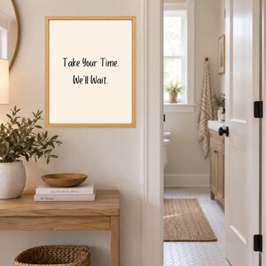 May include: A rectangular wall art piece in a light wood frame featuring a cream-colored background with the black text Take Your Time. We'll Wait. centered in the middle. The artwork is displayed on a wall above a wooden console table.