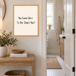 May include: A rectangular wall art piece in a light wood frame featuring a cream-colored background with black text that reads: You Came Here To Pee, Didn't You? The print is displayed on a wall above a wooden console table with books.