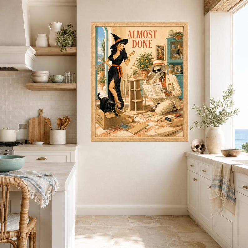 May include: A framed wall art print titled ALMOST DONE featuring a pin-up style witch in a black dress and hat, a skeleton wearing a hat and sunglasses reading instructions, and a black cat in a cardboard box, all in a light wood frame.