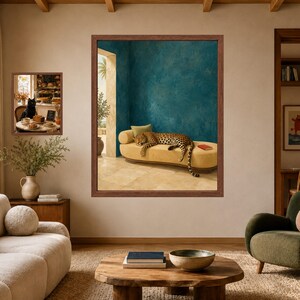 May include: A large framed wall art print featuring a leopard resting on a yellow chaise lounge in a room with teal textured walls and stone flooring. A smaller framed print shows a black cat sitting at a table with food in a bakery setting.