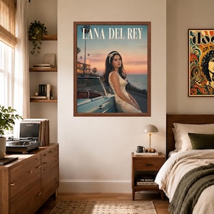 May include: A framed wall poster featuring Lana Del Rey in a white dress leaning against a light blue vintage convertible car at sunset by the ocean. The text LANA DEL REY is printed in white at the top of the image. The poster is displayed on a wall.