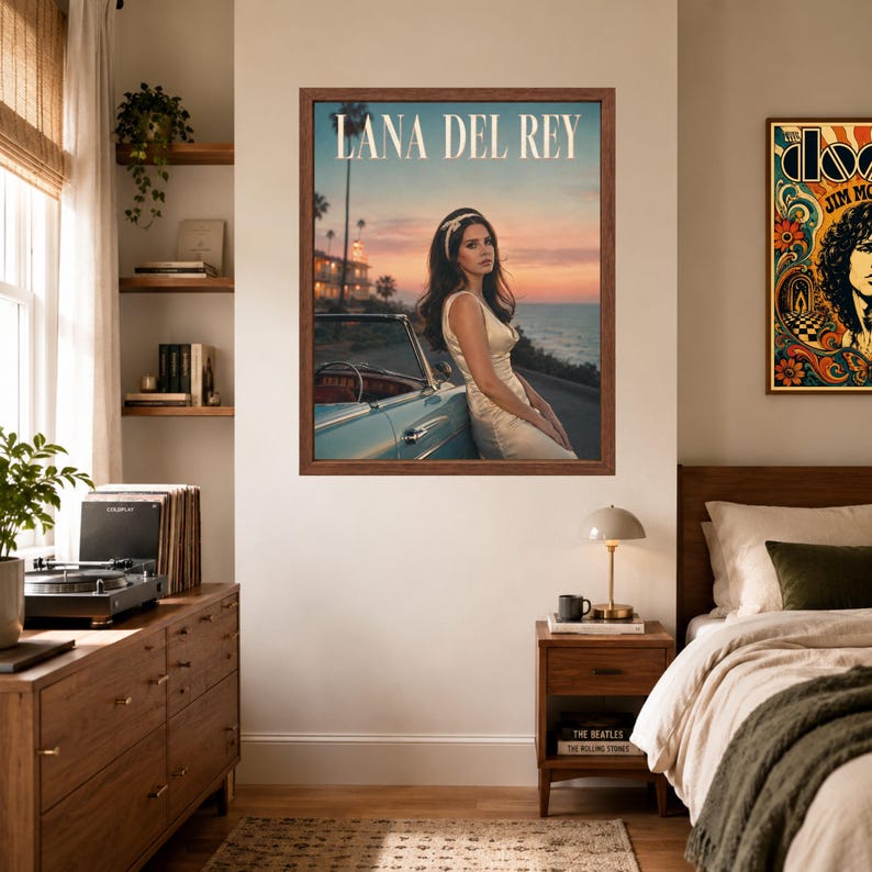May include: A framed wall poster featuring Lana Del Rey in a white dress leaning against a light blue vintage convertible car at sunset by the ocean. The text LANA DEL REY is printed in white at the top of the image. The poster is displayed on a wall.