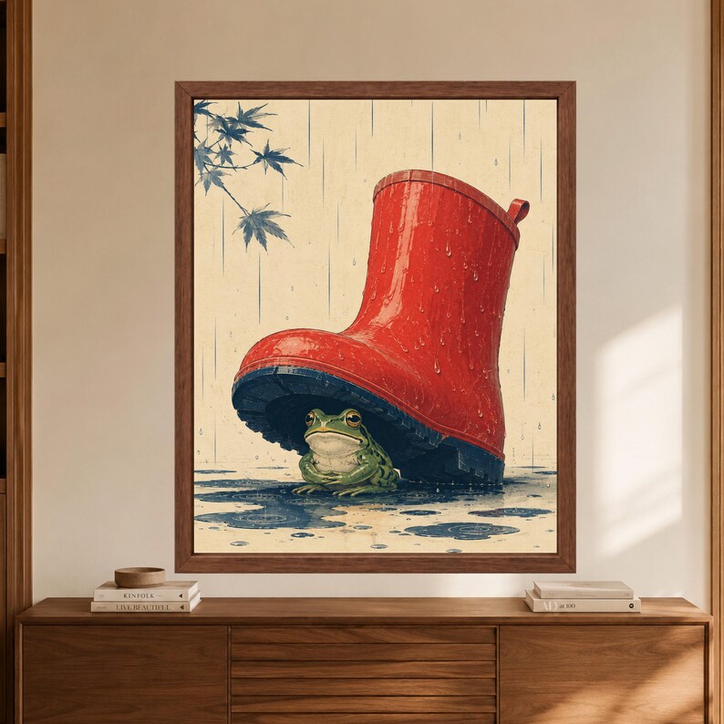 Framed frog wall art showing a green frog sheltering beneath an oversized red rain boot, with falling rain, shallow puddles and blue-green leaves on a warm cream background.