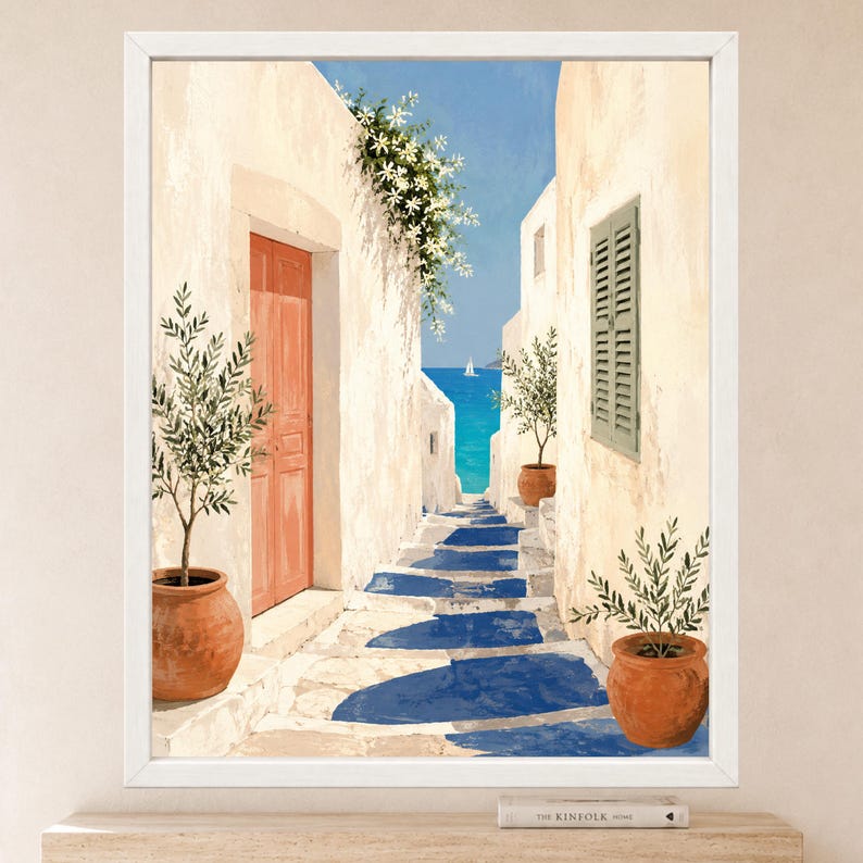 May include: A framed wall art print depicting a Mediterranean alleyway with white stone walls, a coral-colored door, and green potted olive trees. The path leads toward a bright blue sea with a small sailboat under a clear sky.