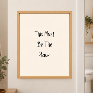 Framed minimalist bathroom wall art with the handwritten black phrase “This Must Be The Place.” stacked vertically on a warm off-white background in a light wood frame beside a bathroom doorway.