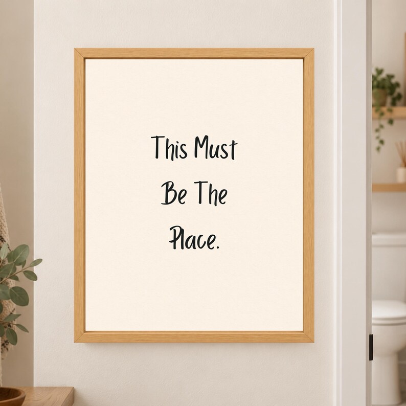 Framed minimalist bathroom wall art with the handwritten black phrase “This Must Be The Place.” stacked vertically on a warm off-white background in a light wood frame beside a bathroom doorway.
