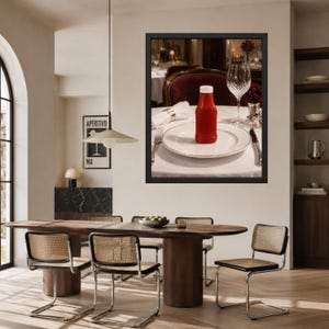 May include: A framed wall art print featuring a red ketchup bottle centered on a white dinner plate. The table setting includes a crystal wine glass, a silver fork, and a silver knife on a white tablecloth, with a dark red chair in the background.