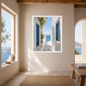 K&ouml;nnte beinhalten: Ein gerahmter Wandkunstdruck, der eine schmale wei&szlig;e Gasse mit leuchtend blauen T&uuml;ren und Fensterl&auml;den zeigt, die den Blick auf das blaue Meer unter klarem Himmel freigibt, inklusive einer Topfpflanze und wei&szlig;en Blumen an einer Mauer.
