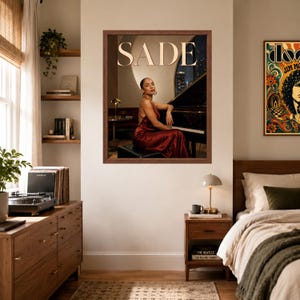 May include: A framed wall poster featuring a portrait of Sade sitting at a grand piano. The singer wears a sleeveless, floor-length, dark red satin dress. The word SADE is printed in large, light-colored capital letters at the top of the image.