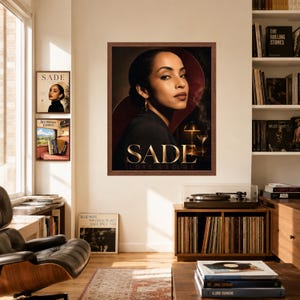 K&ouml;nnte beinhalten: Ein gerahmter Wandkunstdruck mit einem Portr&auml;t der S&auml;ngerin Sade und dem Text SADE LOVE DELUXE. Das Bild zeigt eine dunkelhaarige Person in einem schwarzen Blazer vor einem dunkelroten Hintergrund, mit einem Martiniglas und einer Lampe.
