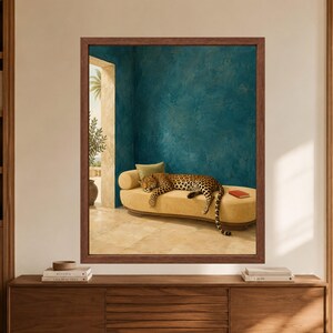 Framed sleeping leopard wall art showing a spotted big cat stretched across a cream curved sofa beneath a deep teal blue wall, with a red book and sunlit Mediterranean terrace in the background.