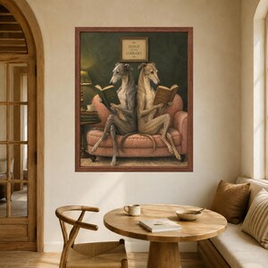 May include: A framed wall art print titled Dogs at the Library featuring two greyhounds sitting back-to-back on a pink sofa, each reading an open book. The background is a dark green wall with a small framed sign above the dogs in a brown frame.