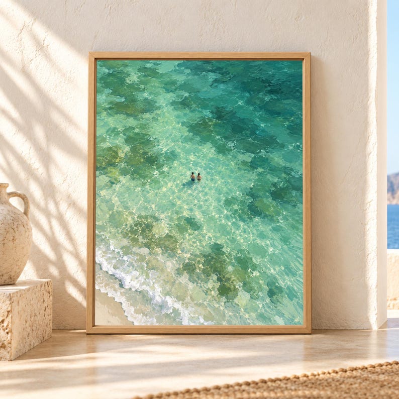 Framed aerial ocean wall art showing two tiny swimmers standing together in clear turquoise and sea green water near a pale sandy shoreline with soft white waves.