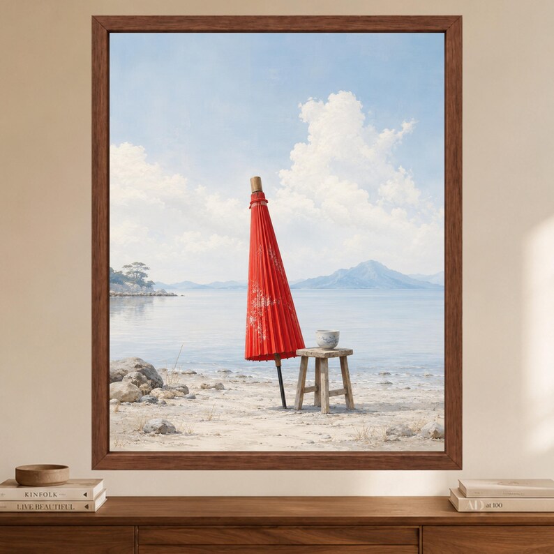 May include: A framed wall art print depicting a closed red Japanese paper umbrella standing upright on a sandy beach next to a small wooden stool with a ceramic bowl on top, set against a calm blue sea and a distant mountain range under a cloudy sky.