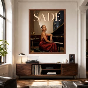 May include: A framed wall art print featuring a portrait of Sade in a dark red satin dress sitting at a black grand piano. The word SADE is printed in large, light-colored capital letters at the top of the image against a warm, dimly lit background.