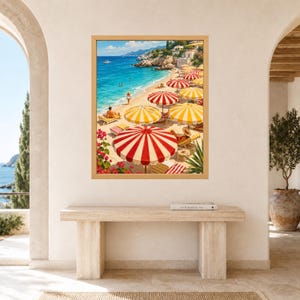 Op de afbeelding: Een ingelijste kunstdruk met een zonnig kuststrandtafereel met turquoise water, rotsachtige kliffen en talloze gestreepte strandparasols in tinten rood, wit en geel. Mensen ontspannen op ligstoelen langs de zanderige kustlijn.