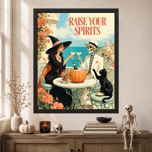 May include: A framed wall art print titled RAISE YOUR SPIRITS featuring a witch in a black dress and hat and a skeleton in a white suit and hat sitting at a table with a pumpkin, wine glasses, and a black cat overlooking a coastal town.